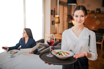 Young and cheerful wautress hold tray with salad bowl and glass of red wine. She look straight on camera. Customer sit behind her and look on phone. She smiles.