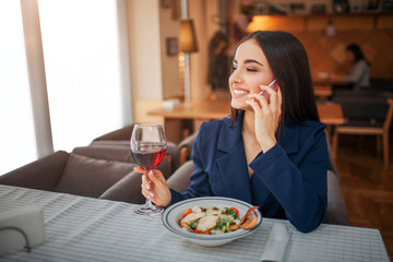 Cheerful young woman sit at table and talk on phone. She smiles. Model hold glass of red wine. Salad bowl stand in front of her at table.