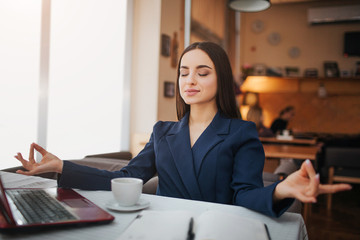 Calm and peaceful young woman sit on table and meditation. She keep eyes closed. Laptop notebook and cup of coffee stand on table.