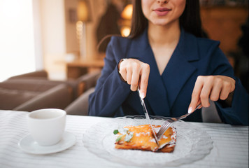 Cut view of nice young woman cut some cake with knife and fork. She sit at table in restaurant. Cup of coffee stand there. She enjoy her meal.