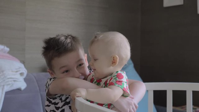 A boy is hugging his baby sister, who is standing in a crib. Both of them are wearing T shirts and obviously enjoying the company of each other