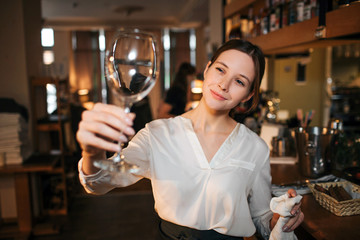 Picture of young waitress stand and look at clean glass for wine. She hold white towel in another hand. Waitress work at bar in restaurant.