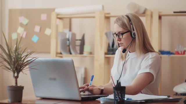 a beautiful young girl works as a call center operator, talking on the headset and working at the computer