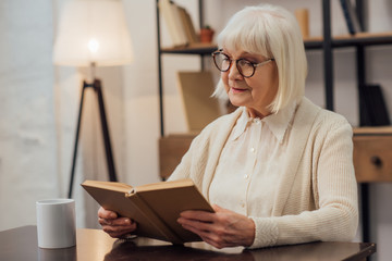thoughtful senior woman in glasses sitting at table and reading book at home