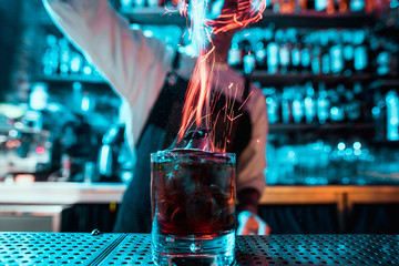 Glass of fiery cocktail on the bar counter against the background of bartenders hands with fire....
