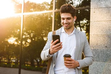 Photo of smiling businessman in suit holding mobile phone, while standing outdoor near building with takeaway coffee