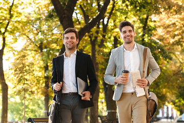 Photo of successful businessmen in suits walking outdoor through green park with takeaway coffee and laptop, during sunny day