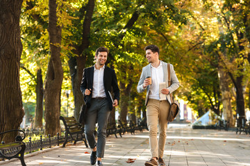 Photo of bearded businessmen in suits walking outdoor through green park with takeaway coffee and...