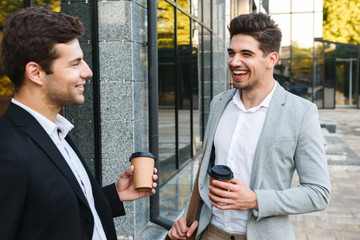 Photo of young entrepreneurs in suits standing outdoor near building with takeaway coffee, during sunny day
