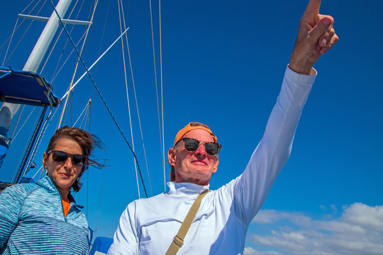 Older Baby Boomer Caucasian Couple With Man Pointing Into The Distance. Deep Blue Sky With Sailboat Rigging In Background