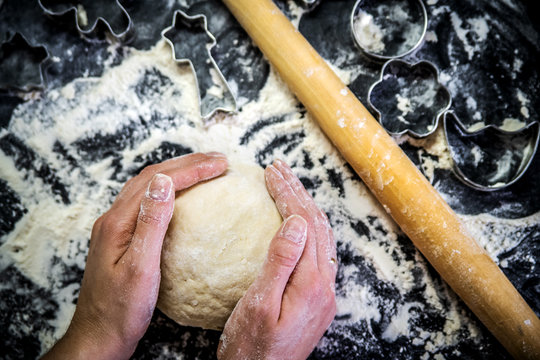 Knead dough in the hands of the cook, rolling pin and cookie cutters on the table covered with flour.