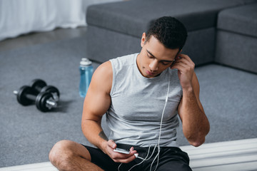 Fototapeta na wymiar selective focus of bi-racial man using smartphone and listening music while sitting on fitness mat