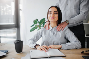 Young woman get attention from boss. He does shoulder massage for her. She is relaxed and pleased. Young woman sit in room at table. Guy stand behind her.