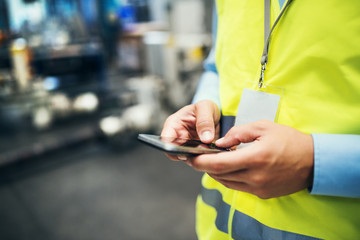 An unrecognizable industrial man engineer with name tag in a factory, using smartphone.
