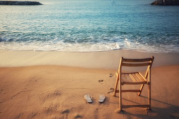 White sandals and wooden chairs placed on the sand with blue waves ocean.