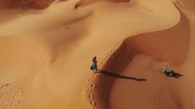 AERIAL. Woman weared in long dress walking in the Dubai desert sand dunes with footsteps in the sand during sunset