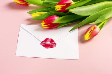 Valentine's day background. Love letter concept. White envelope with red lipstick kiss and bouquet of tulips on pink pastel.