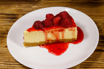 Piece of tasty New York cheesecake with raspberries and raspberry jam in a white plate on wooden table