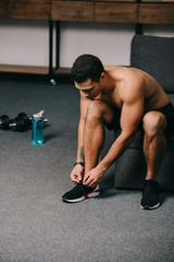 strong bi-racial man tying shoelaces near dumbbells and sport bottle in living room