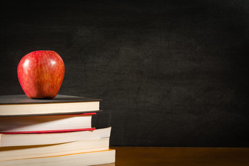 Stack of books and a red apple on a desk front of a blank chalkboard