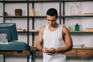 handsome strong bi-racial man using smartphone in living room near treadmill