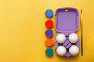 Easter composition. Set of four white eggs in a purple box, multi-colored jars of paint and a brush for drawing on a yellow background