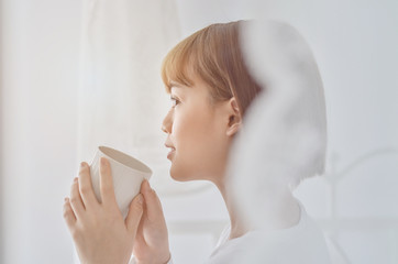 Beautiful woman holding a white cup of coffee in the morning bedroom.Gray bedroom, she is drinking coffee.
