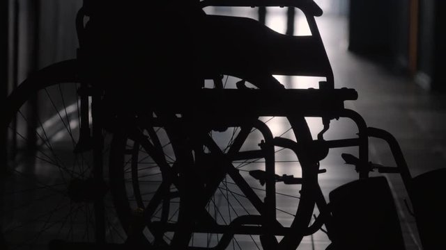 Tilt up shot of silhouette of man with stick limping to wheelchair along dark corridor in hospital