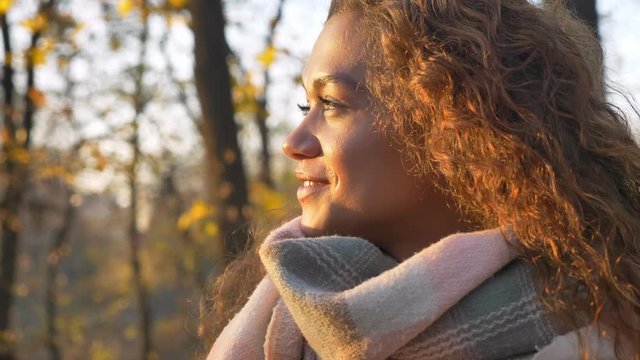 Portrait of caucasian curly-haired woman smilingly watching into camera and turning to sun on autumnal park background.