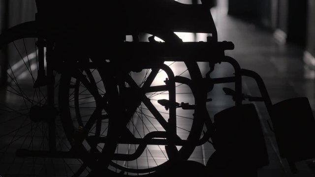 Tilt up shot of silhouettes of man supporting woman limping with stick along hallway of hospital; wheelchair in foreground