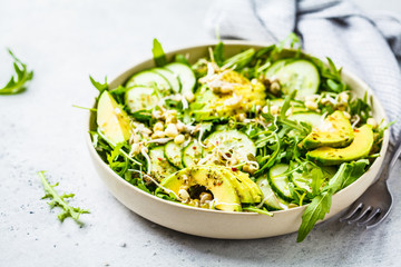 Healthy green salad with avocado, cucumber and arugula in white dish.