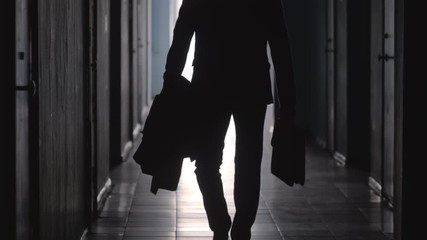 Tilt up shot of silhouette of unrecognizable businessman wearing formal suit and carrying coat and briefcase walking away from the camera along hallway in office center - Powered by Adobe