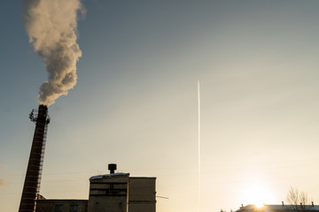 At the setting sun, the plane flies against the background of the old factory where the pipes smoke. Air pollution in the city