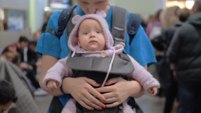 Mother carrying baby daughter in kangaroo carrier. They waiting at the airport. Woman kissing lovely quiet child in knitted pink hoodie onesie
