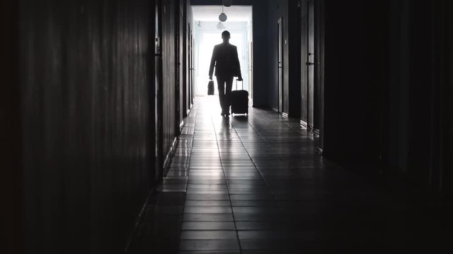 Full length shot of silhouette of unrecognizable businessman walking with luggage and briefcase towards the camera along hallway in hotel