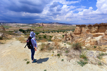 A young girl stands on the edge of a cliff in Cappadocia and admires the surrounding space against the backdrop of a stormy cloudy sky and mountain landscape.