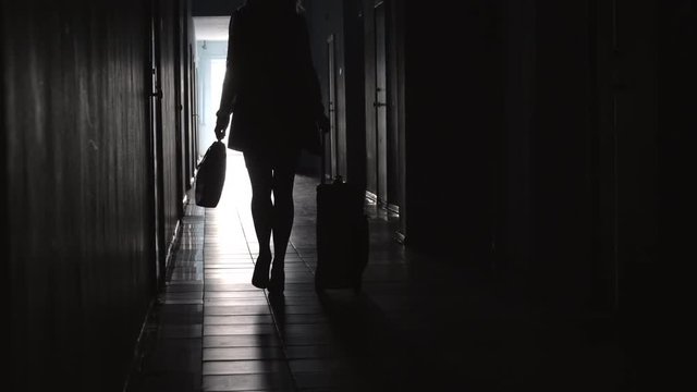 Tilt up rear view of silhouette of unrecognizable business lady carrying briefcase and pulling luggage while walking along hallway in hotel
