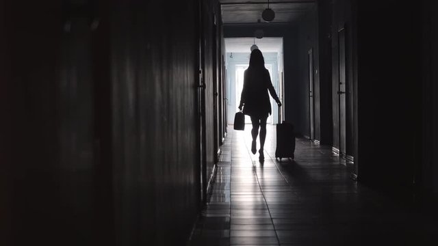 Full length silhouette of unrecognizable businesswoman carrying briefcase and pulling luggage while walking along hallway in hotel