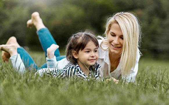 Lovely happy little girl lying on the green grass and playing with her beautiful mother in the park. Woman and her little kid daughter enjoy the holiday. Mom and child has fun outdoors. Mother's day