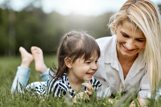 Image of happy little girl lying on the green grass and playing with her beautiful mother in the park. Woman and her little daughter enjoy the holiday. Mom and child has fun outdoors. Mother's day