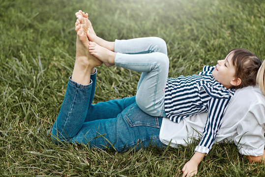 Horizontal side view of beautiful woman lying and playing with her cute little girl on the green grass outdoors. Happy child and mother spend time together in the park. Mother's day concept