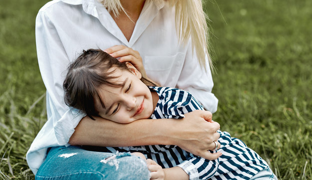 Lovely image of cute little child girl lying on her mother's legs outside. Loving mother and daughter sitting on the green grass in the park. Mom and kid cuddling. Good relationship. Mother's day