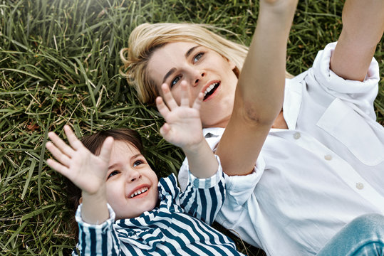 Overhead image of pretty blonde woman playing with her cute little child, lying on green grass outdoor. Loving mother and daughter spend time together in the park. Mom and kid has fun. Mother's day