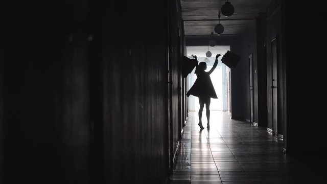 Silhouette of joyous woman carrying shopping bags, raising arms up, spinning around and walking along hallway in hotel 