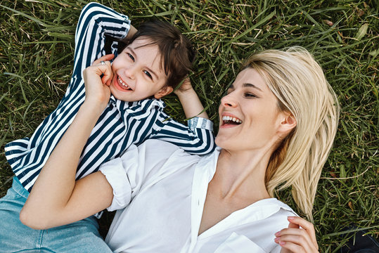 Top view of happy blonde woman smiling and playing with her cute little child, lying on green grass outdoor.Loving mother and daughter spend time together in a park. Mom and kid has fun. Mother's day