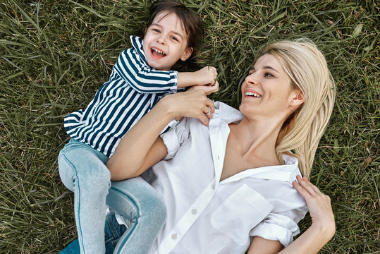 Overhead view of happy blonde woman playing with her cute little child, lying on green grass outdoor. Loving mother and daughter spend time together in the park. Mom and kid has fun. Mother's day