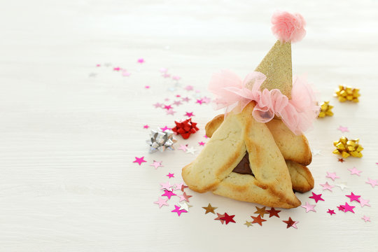 Purim celebration concept (jewish carnival holiday). Traditional hamantaschen cookies with cute carnival hat over white wooden table.