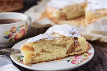 A piece of delicious apple charlotte sponge cake next to a cup of flavored black tea next to a whole cake on a wooden table