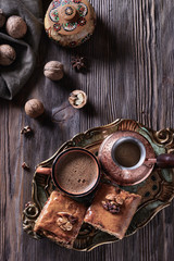 A cup of freshly brewed coffee next to a cezve and baklava on a wooden vintage tray and on a wooden background next to nuts, top view, vertical
