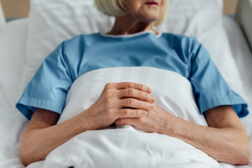 cropped view of senior woman with folded hands lying in bed in hospital